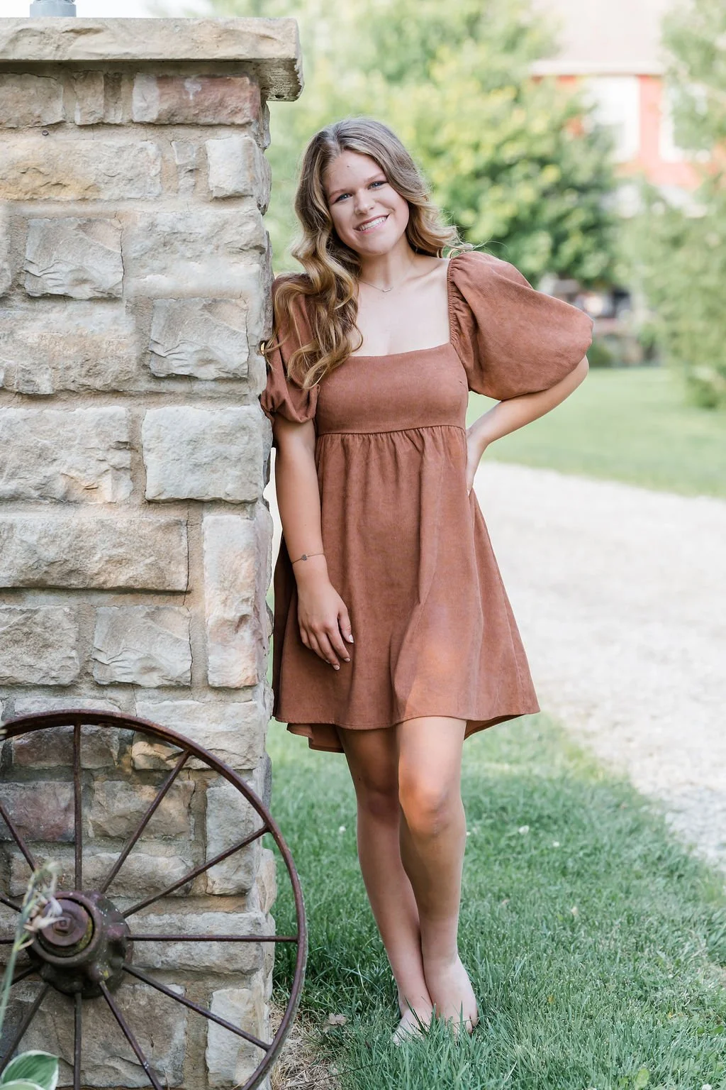 A young woman with long, wavy, light brown hair, smiling and posing outdoors next to a stone wall. She is wearing a brown dress with puffed short sleeves and stands on grass with a blurred background of trees and a pathway.