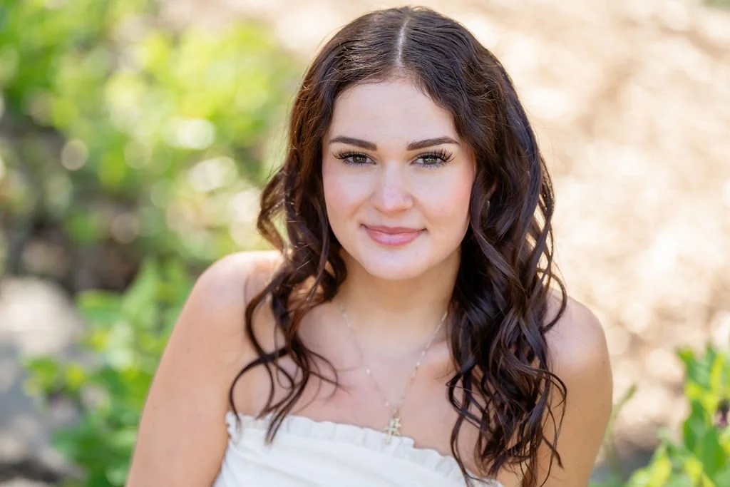 Young woman with long, curly brown hair and light makeup outdoors, smiling gently at the camera.