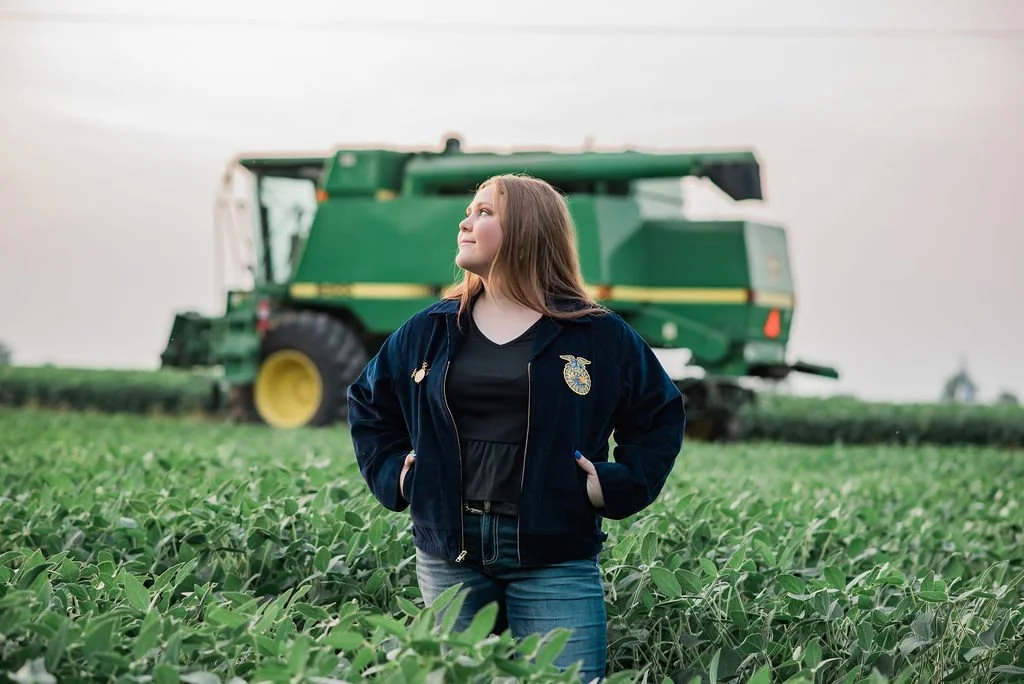 A young woman standing in a soybean field with a large green combine harvester in the background, wearing a navy blue jacket with a USDA emblem.
