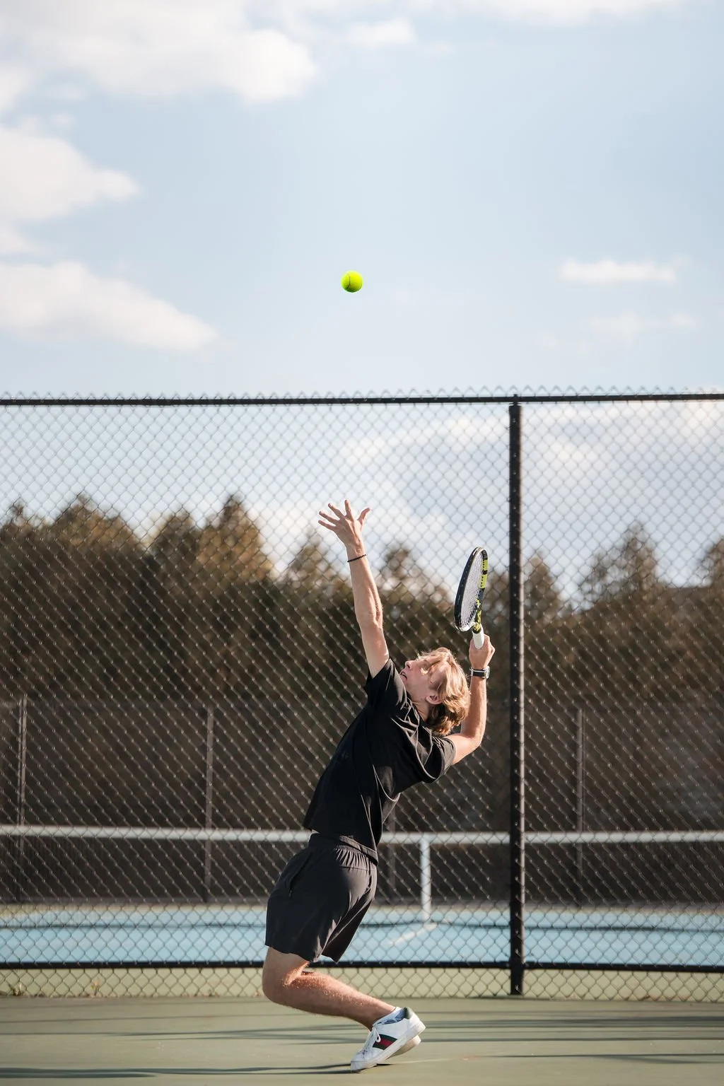 A young man playing tennis on an outdoor court, preparing to hit a tennis ball with his racket. The sky is partly cloudy, and there is a chain-link fence surrounding the court, with trees in the background.