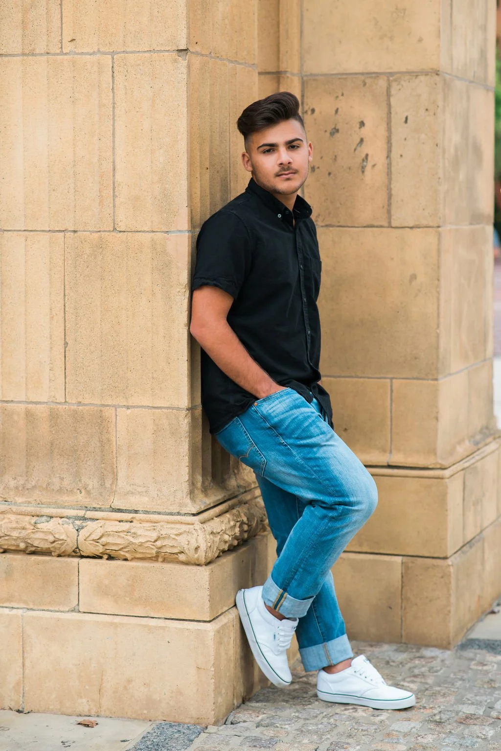 A young man in a black shirt and blue jeans leaning against a light-colored stone wall, with one hand in his pocket and the other hanging by his side, looking at the camera.