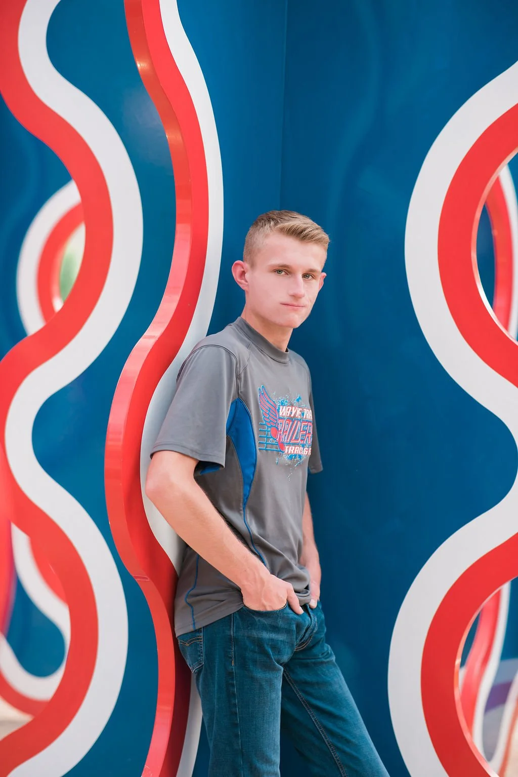 A young man with short blond hair wearing a gray and blue T-shirt and blue jeans, standing with his hands in his pockets against a colorful, curvy wall with red, white, and blue stripes.