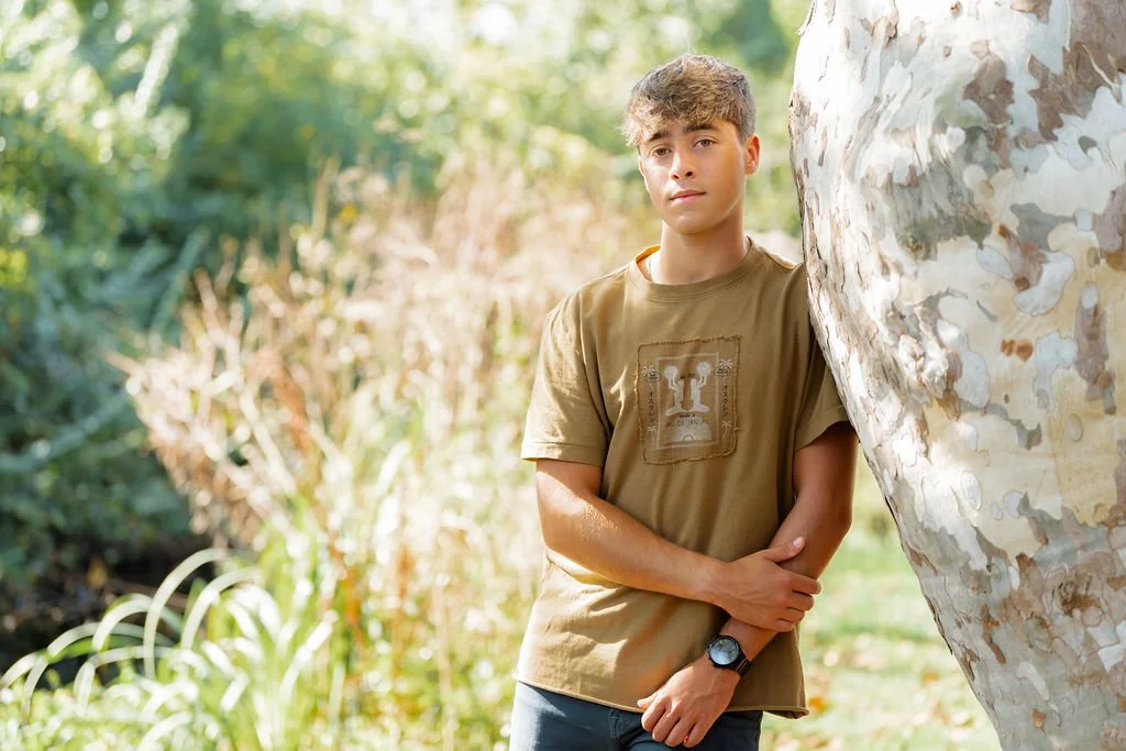 A teenage boy with light brown hair wearing a tan T-shirt and wristwatch, standing outdoors beside a large tree with a mottled bark, in a natural setting with sunlight and greenery.