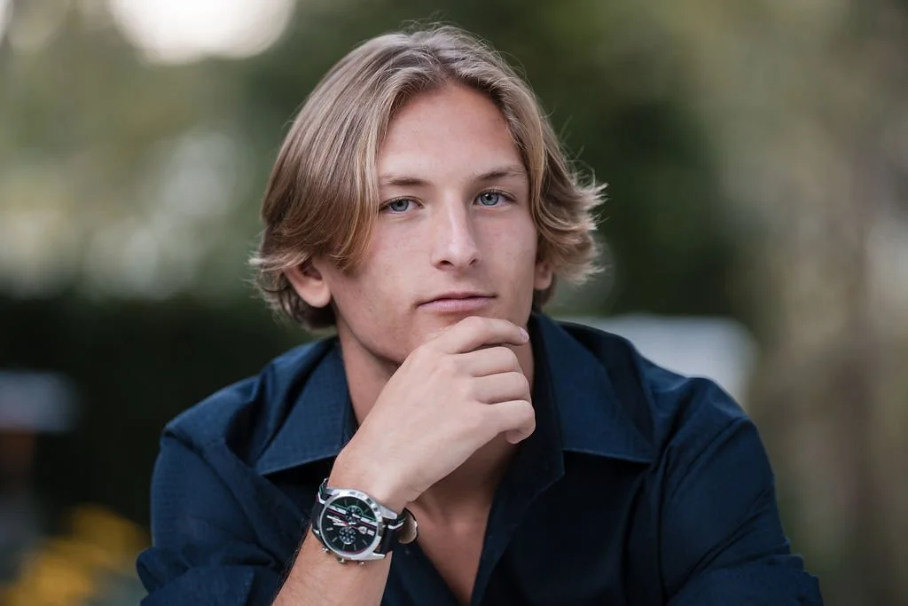Young man with blond hair, blue eyes, wearing a dark blue shirt and a wristwatch, resting his chin on his hand outdoors with blurred greenery in the background.