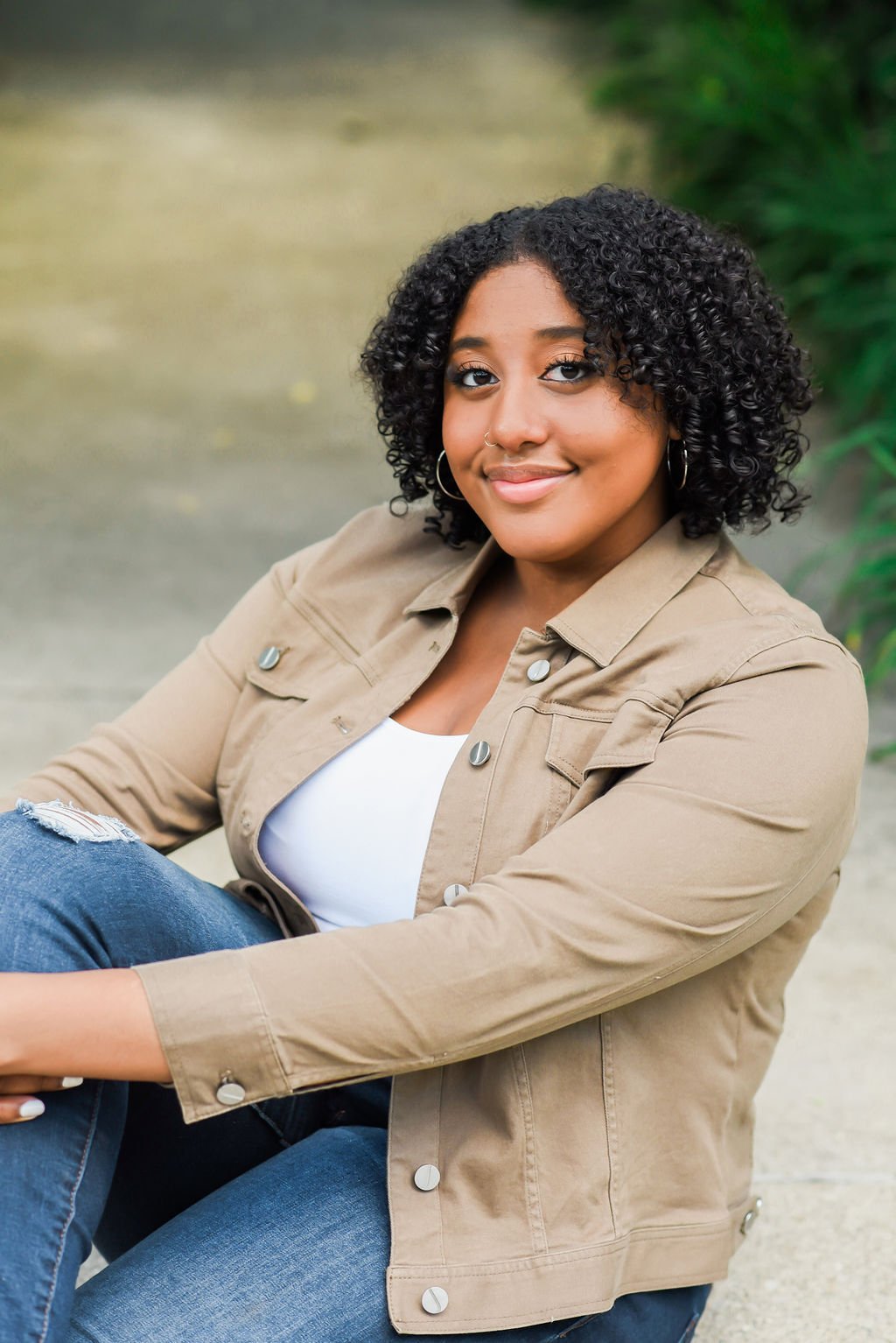 A young woman with curly black hair, light brown skin, wearing earrings, a nose ring, a white top, a light brown jacket, and ripped jeans, sitting outdoors near green foliage, smiling at the camera.