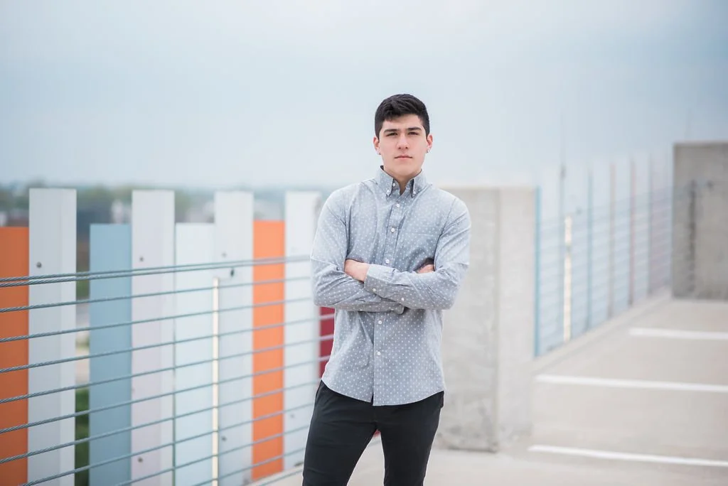 A young man with dark hair standing on a rooftop parking lot with his arms crossed. He's wearing a light gray, button-up shirt and black pants, with a colorful abstract fence and a cloudy sky in the background.