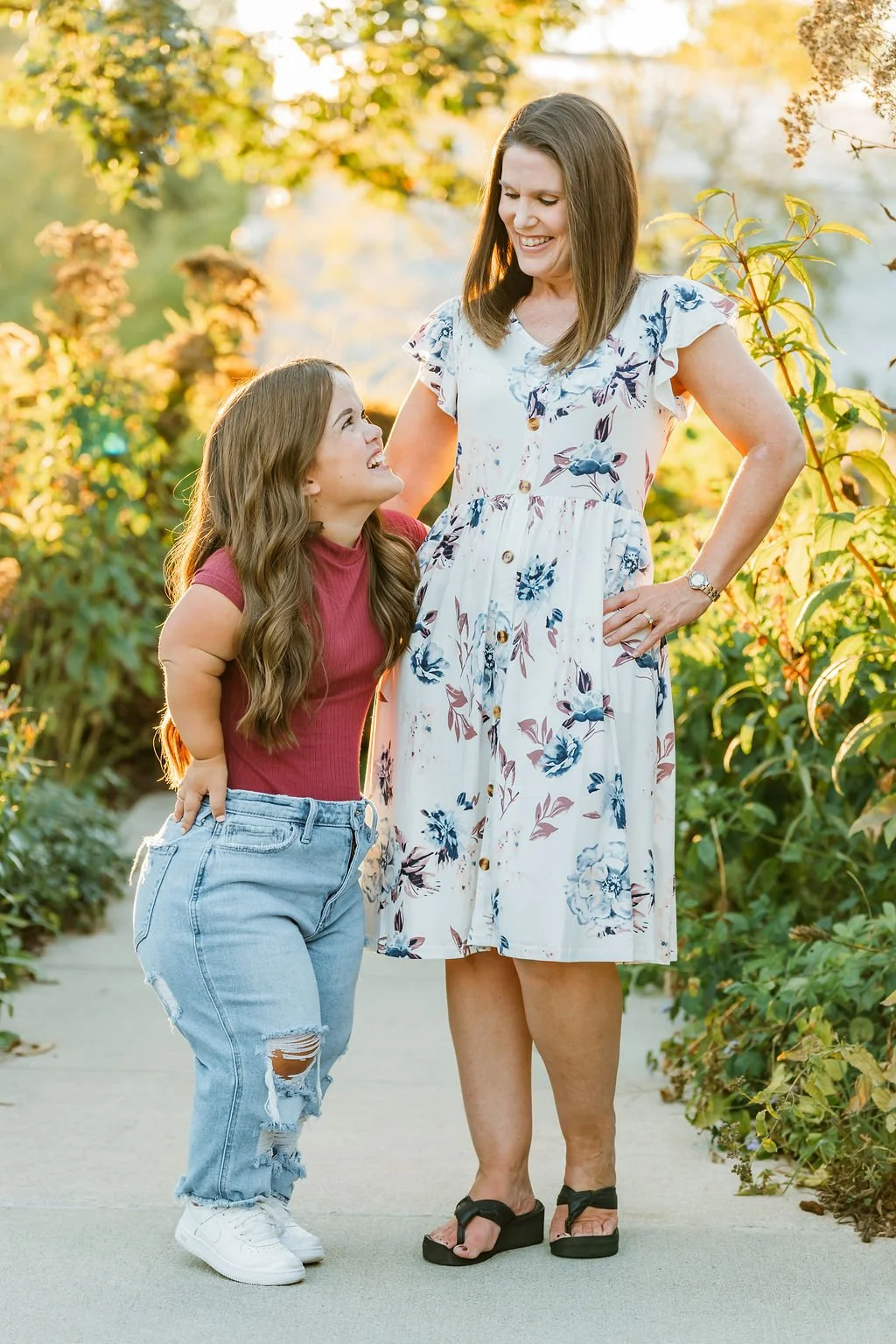 A woman and a young girl are outdoors in a garden with green foliage and flowers in the background, smiling at each other.