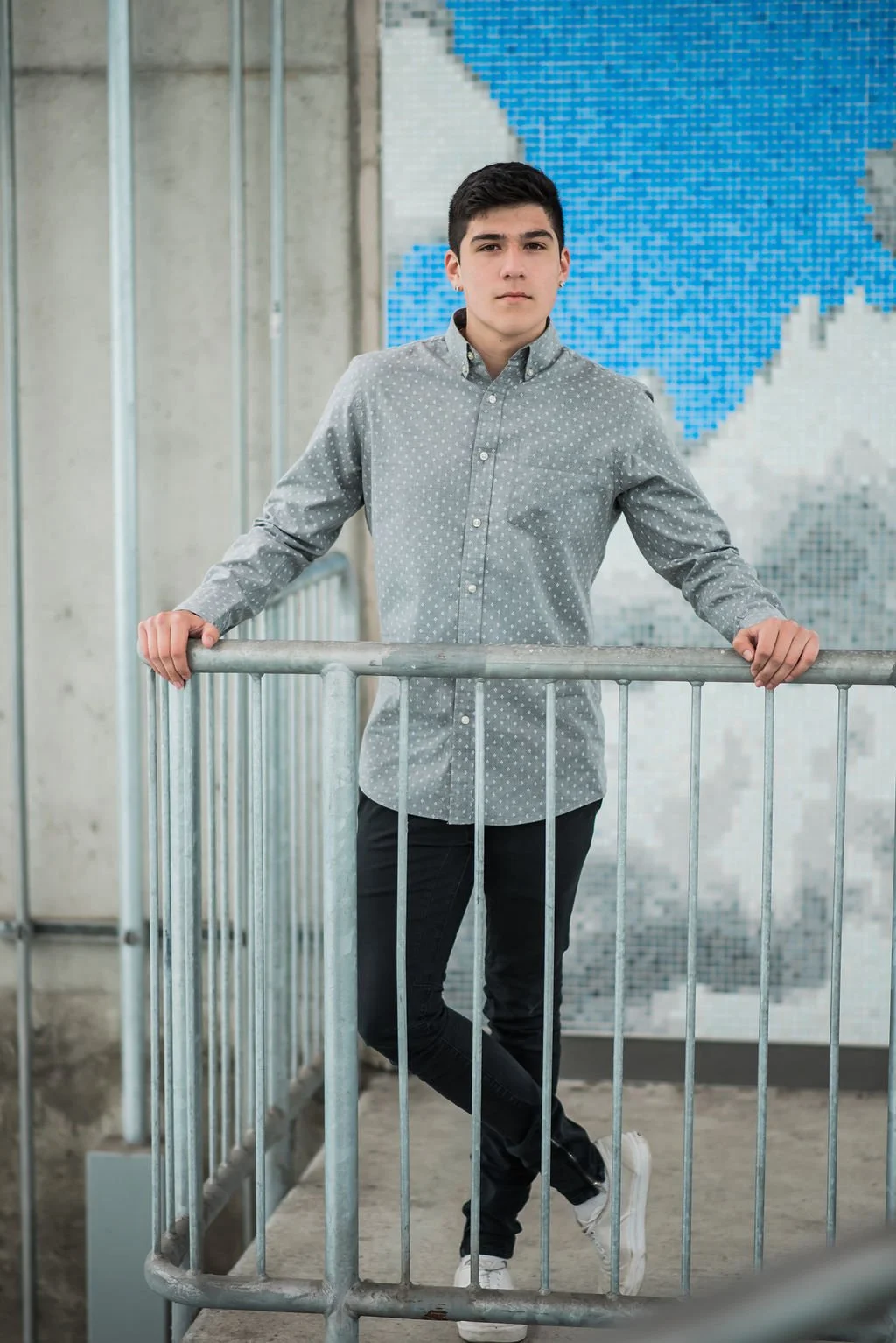 Young man standing behind a metal railing in an indoor space with a large abstract blue and gray pattern on the wall behind him.