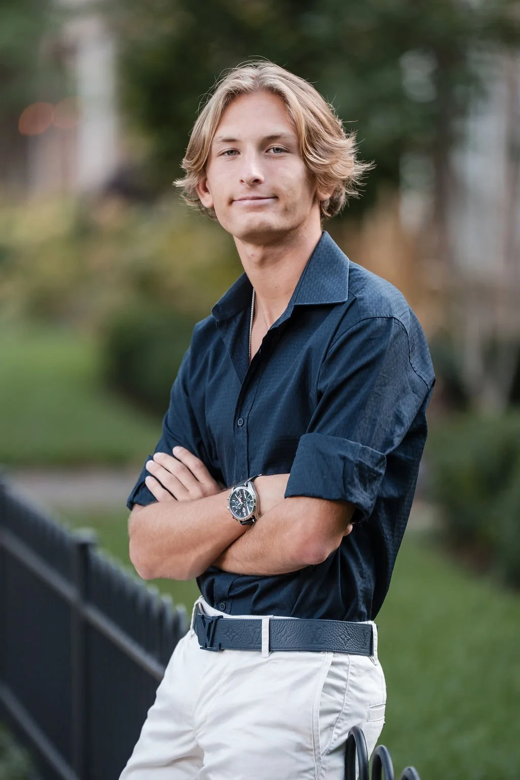 A young man with blonde hair and fair skin, wearing a navy blue button-up shirt with the sleeves rolled up, and beige pants with a black belt. He stands outdoors in front of a blurred background of greenery, with arms crossed and a slight smile.