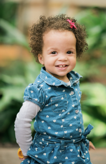 A young girl with curly hair, a pink hair clip, and a big smile. She wears a blue dress with white hearts and a white long-sleeve shirt underneath. She is outdoors with green foliage in the background.
