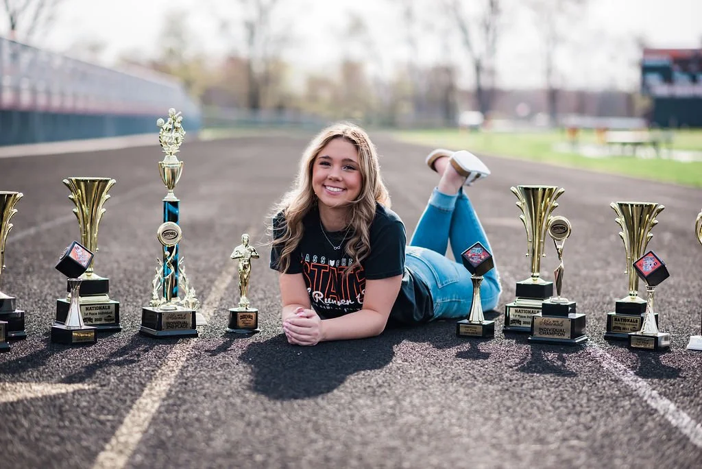 Young girl lying on her stomach on a running track, smiling, surrounded by multiple trophies and awards.