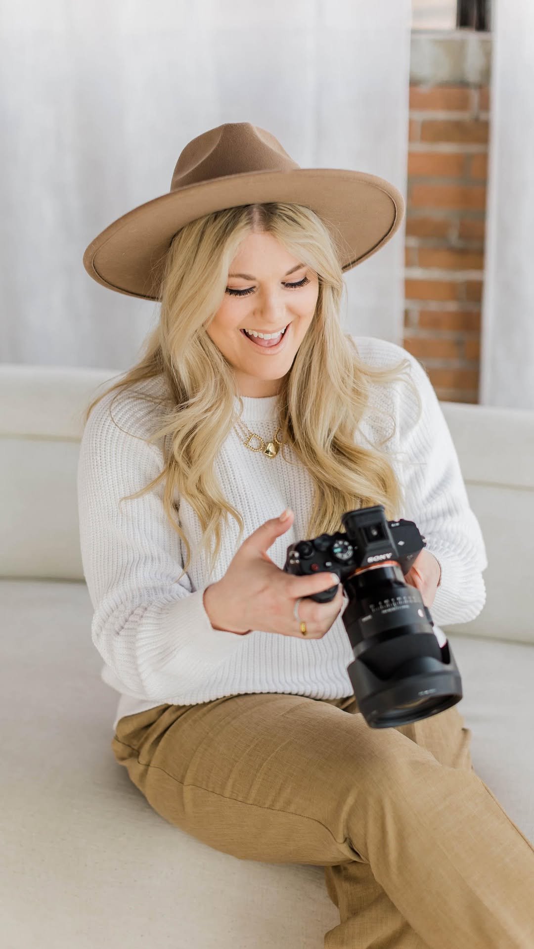 A woman with long blonde hair, wearing a wide-brimmed tan hat, a white sweater, and tan pants, is sitting on a couch, looking at a camera she is holding, smiling.