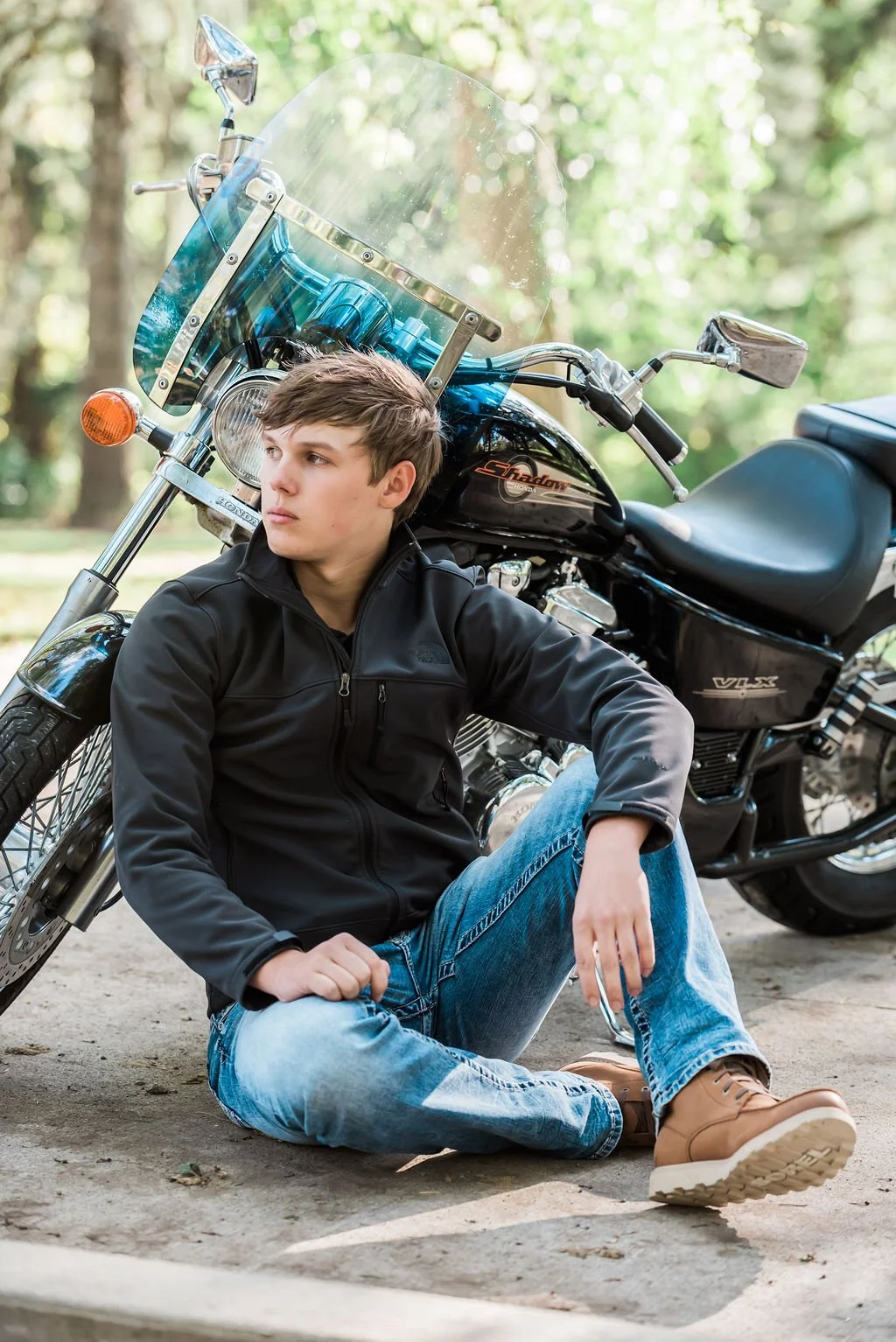 A young man with light brown hair in a black jacket and jeans sits on the ground next to a black motorcycle with a clear windshield in a wooded area.