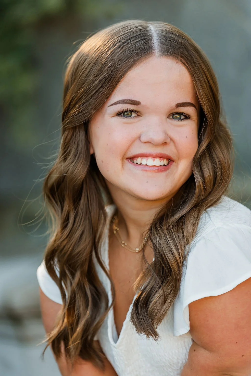 Close-up portrait of a smiling woman with long, wavy brown hair, wearing a white top and a layered gold necklace, outdoors with a blurred natural background.