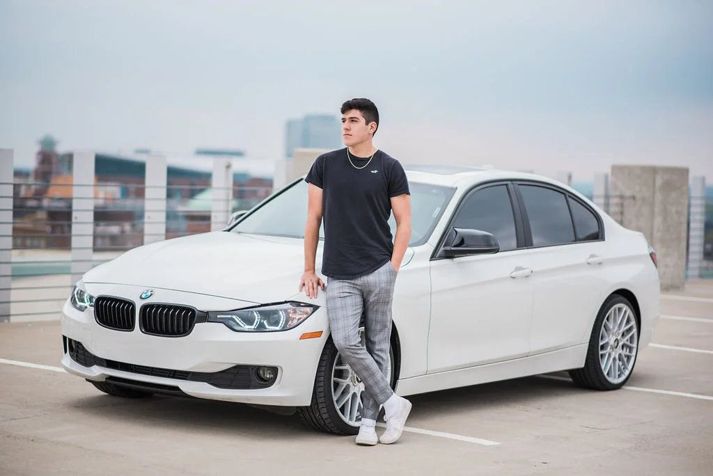 A young man in a black t-shirt and plaid pants leaning against a white BMW sedan in a parking lot.