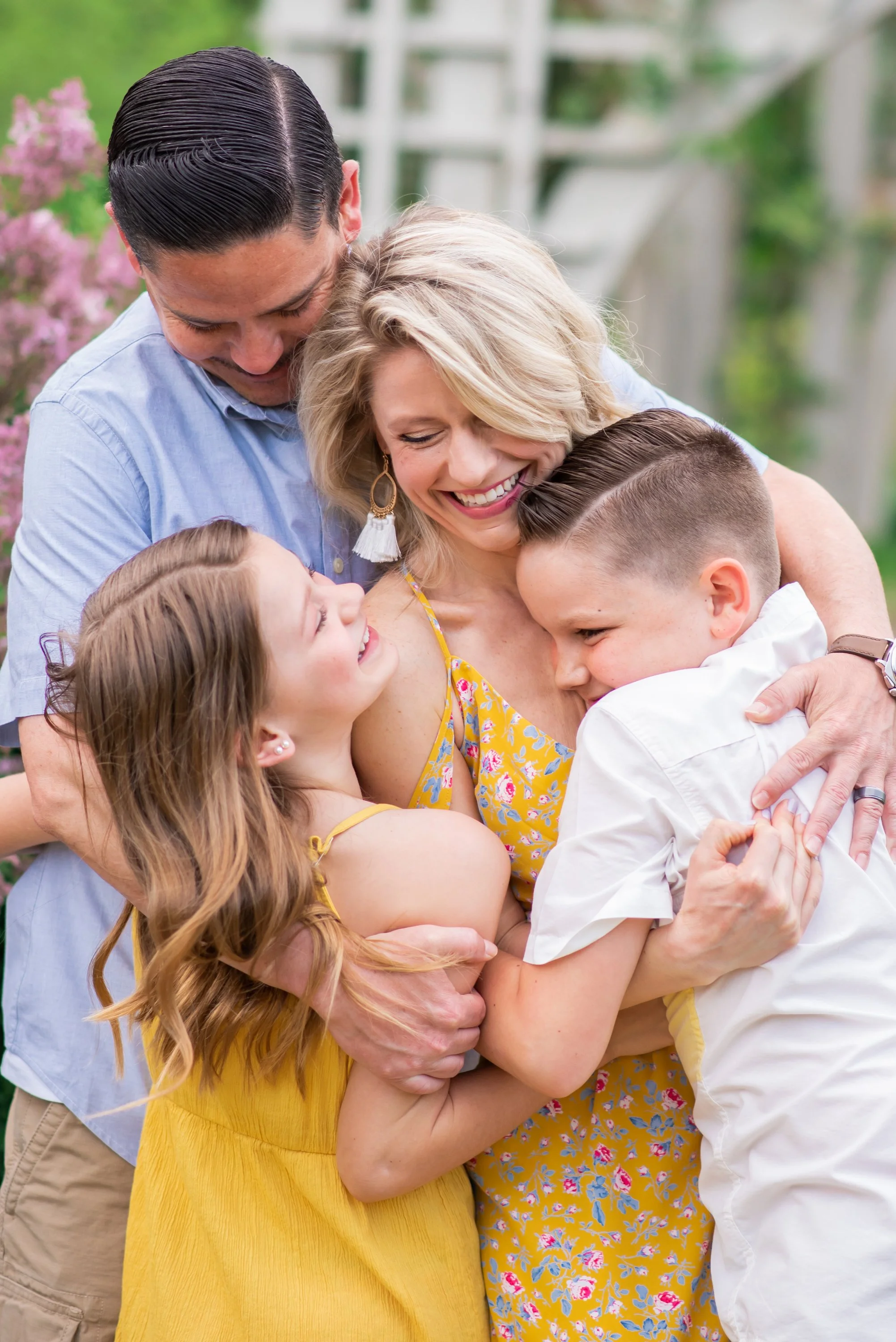 A family of five hugging and smiling outdoors, surrounded by greenery and pink flowers, showing love and joy.