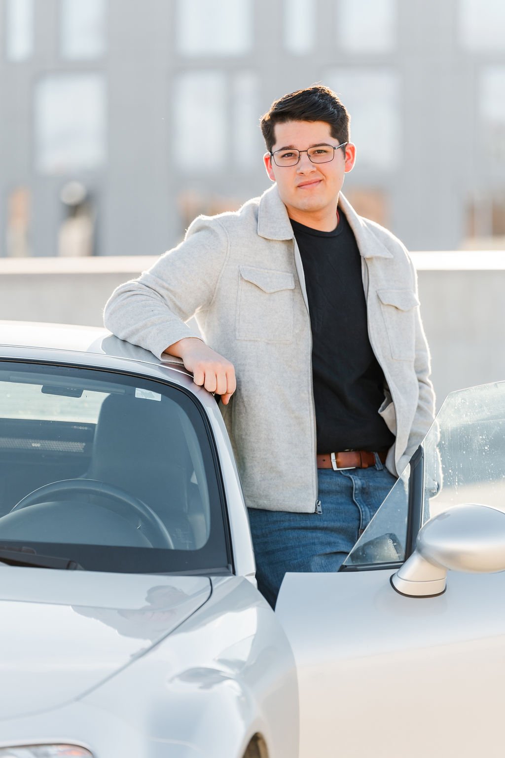 Young man with glasses, wearing a black shirt and gray jacket, standing next to a silver sports car.