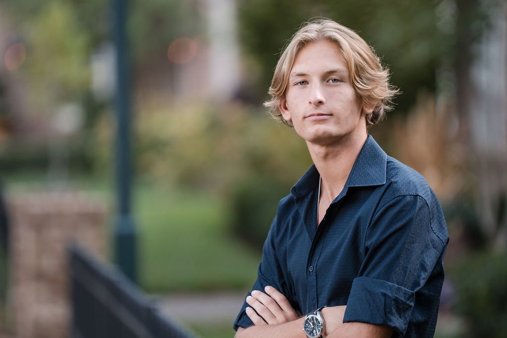 A young man with blonde hair wearing a dark blue shirt and a watch, standing outdoors with arms crossed, in a park or garden setting.