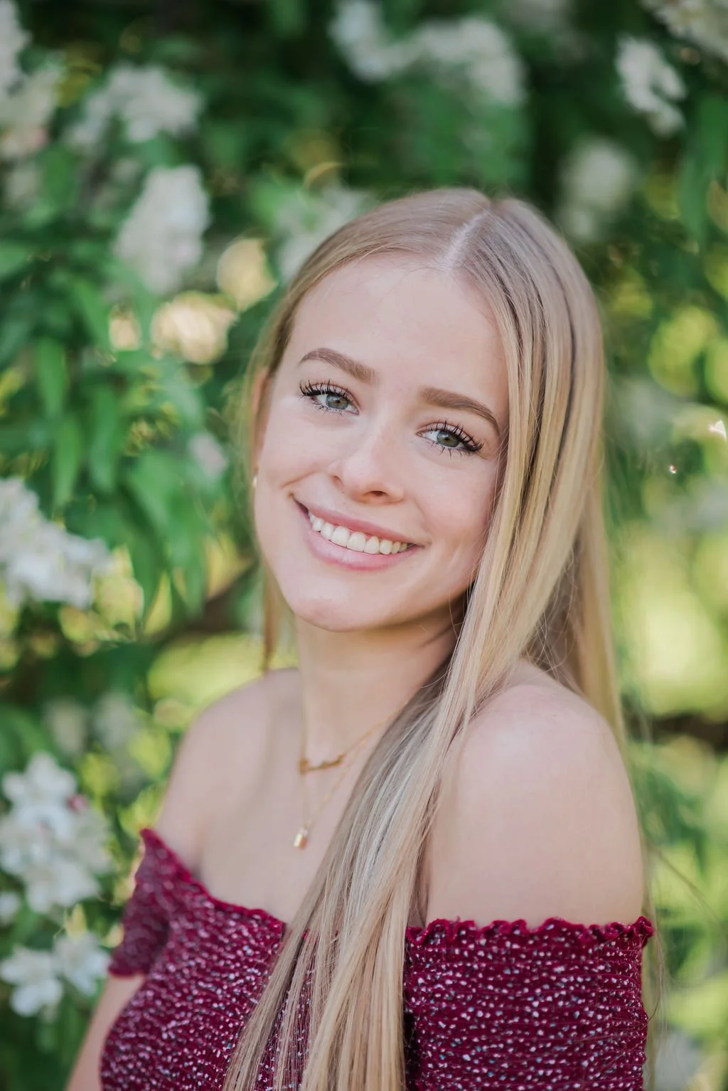 A young woman with long blonde hair smiling outdoors among green foliage and white flowers.
