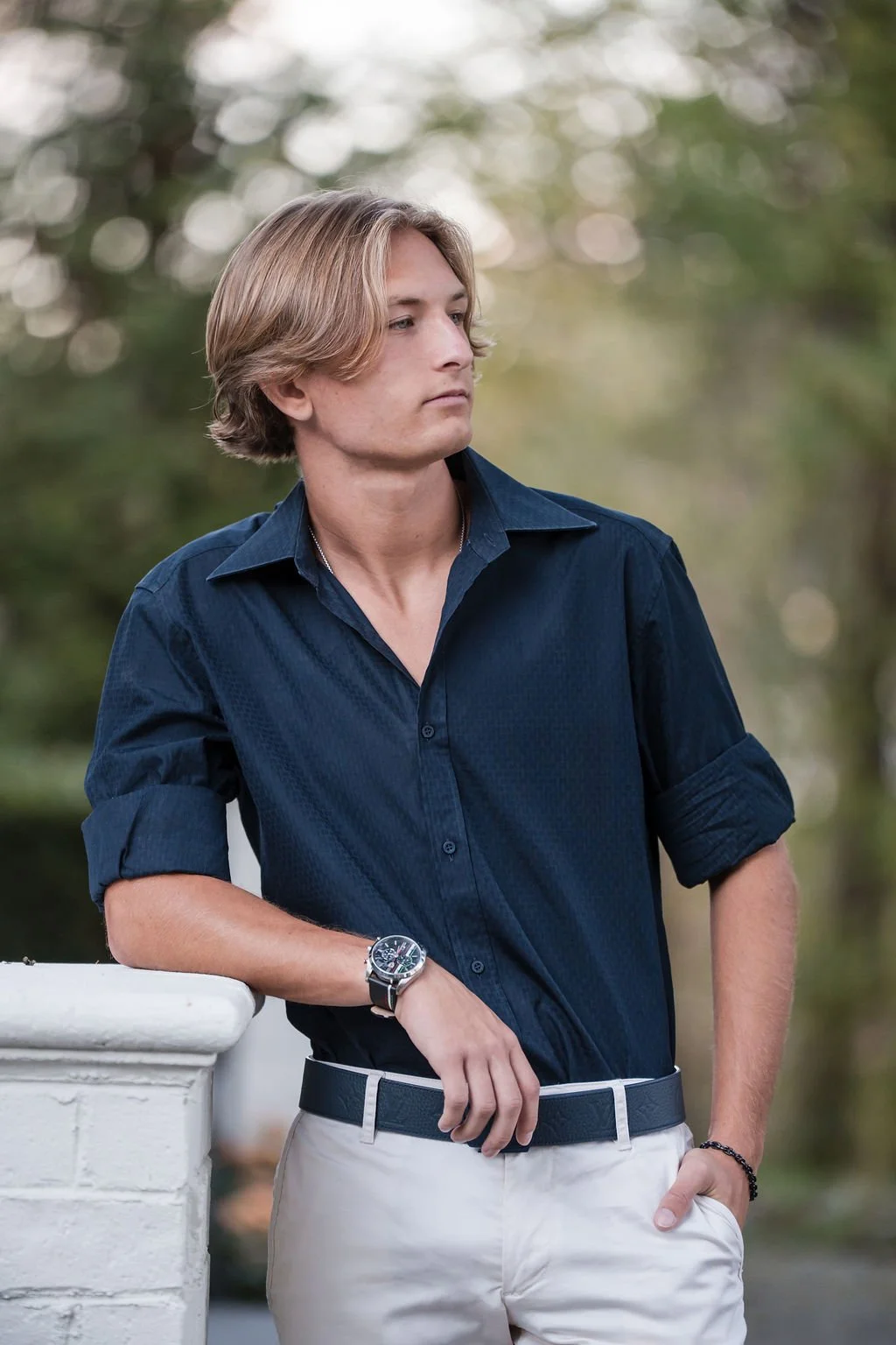 A young man with blond hair wearing a dark blue shirt and beige pants, leaning against a white railing outdoors with a blurred natural background.
