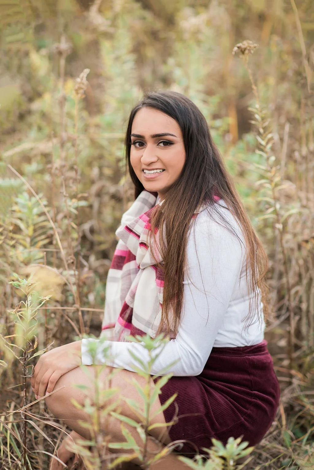 A young woman with long dark hair, smiling, squatting among tall grass and plants outdoors in a natural setting with a blurred background.