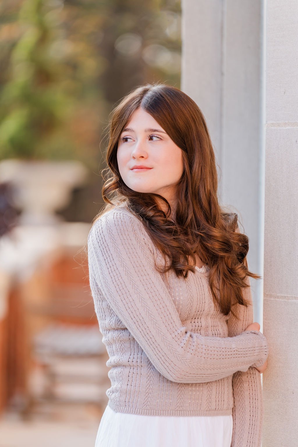 A young woman with long brown hair stands outdoors near a stone wall, looking to the side with a thoughtful expression.