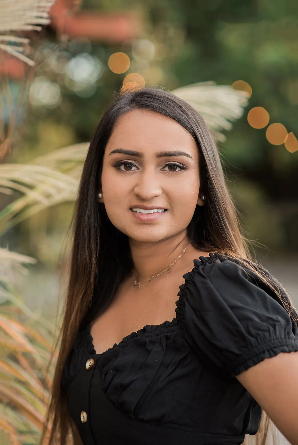 A young woman with long brown hair, wearing a black top with ruffled sleeves, earrings, and a necklace, standing outdoors with blurred greenery and lights in the background.
