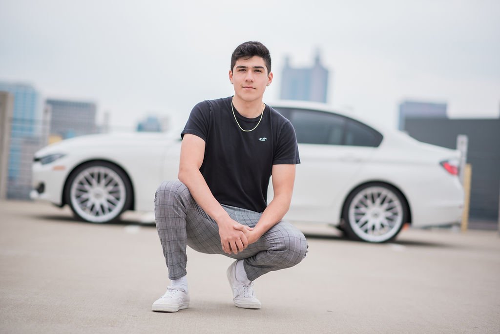 A young man is squatting in front of a white luxury car on a rooftop parking lot with a city skyline in the background.