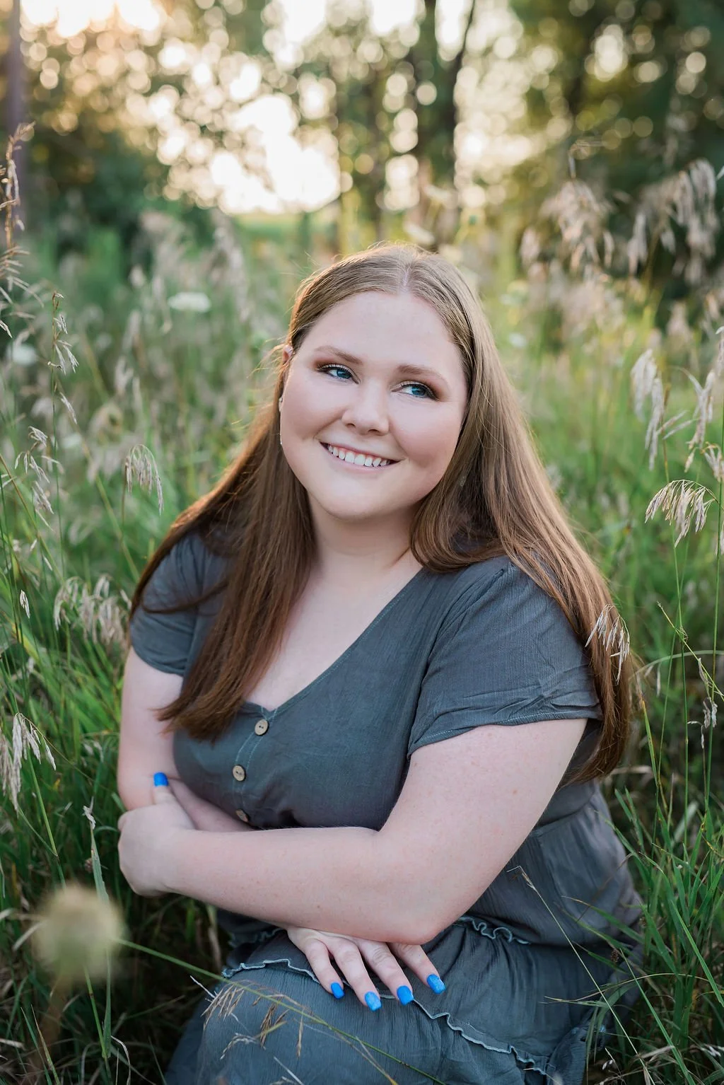 A smiling woman with long brown hair and blue eyes sitting outdoors in a field of tall grass, with trees and sunlight in the background.