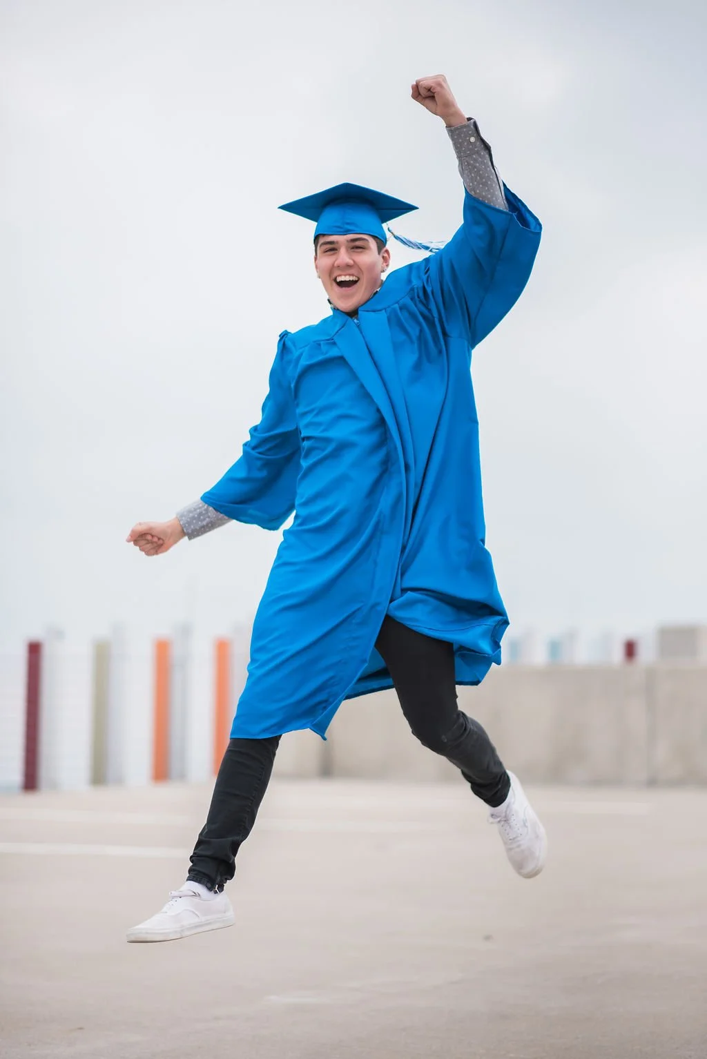 Young man in blue graduation gown and cap celebrating with a joyful expression, jumping in an outdoor space.