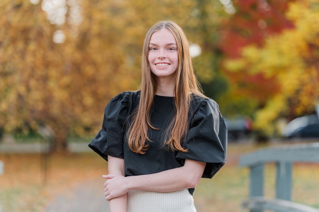 Young woman with long red hair smiling outdoors on a fall day, wearing a black puff-shoulder top and a light-colored skirt.