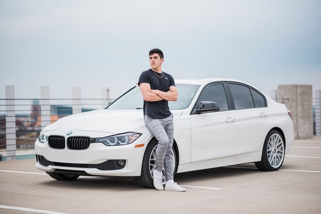 A young man standing with arms crossed in front of a white BMW parked on a rooftop parking deck during daytime.