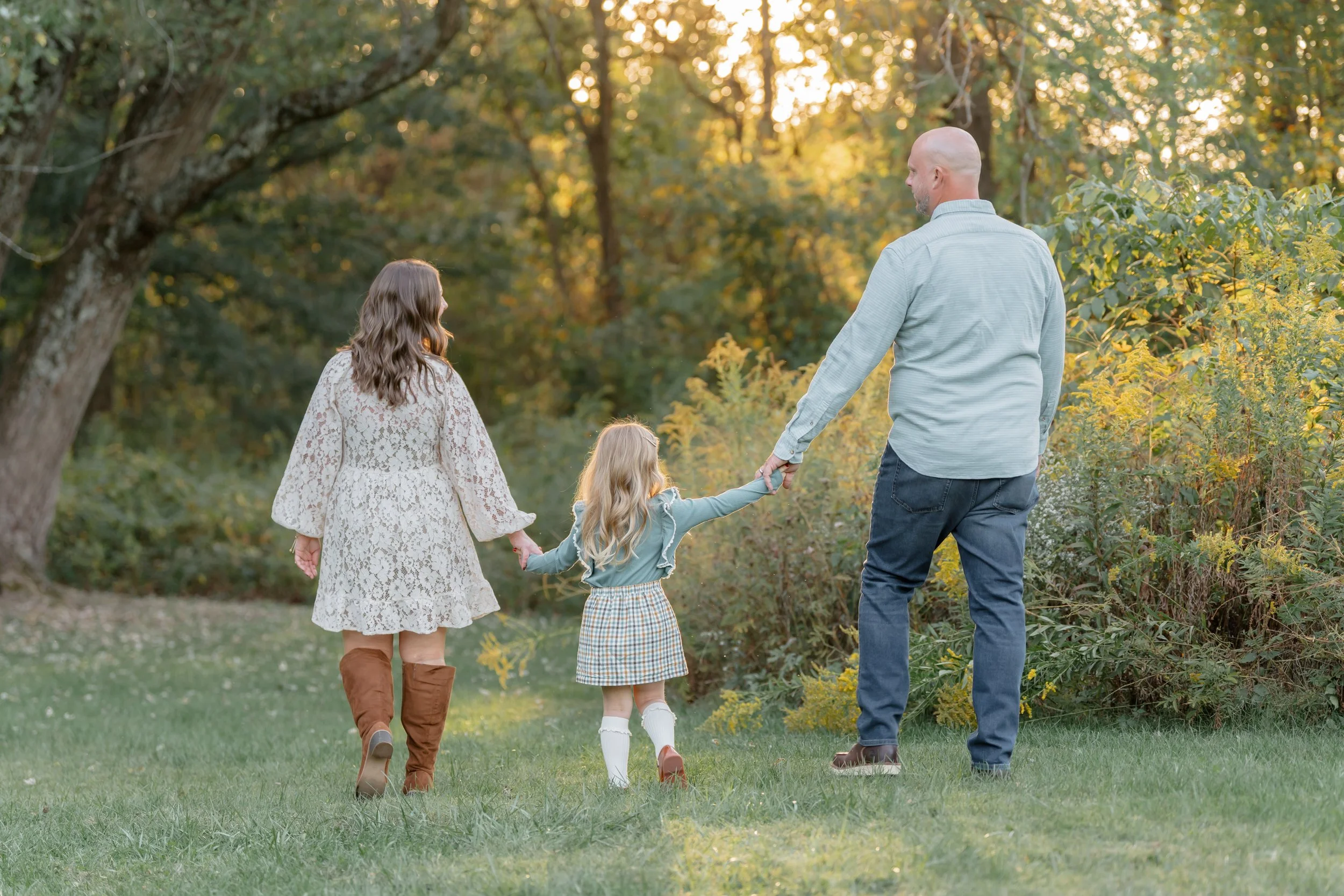 A family of three walking outdoors in a park during autumn, holding hands, with trees and foliage in the background.