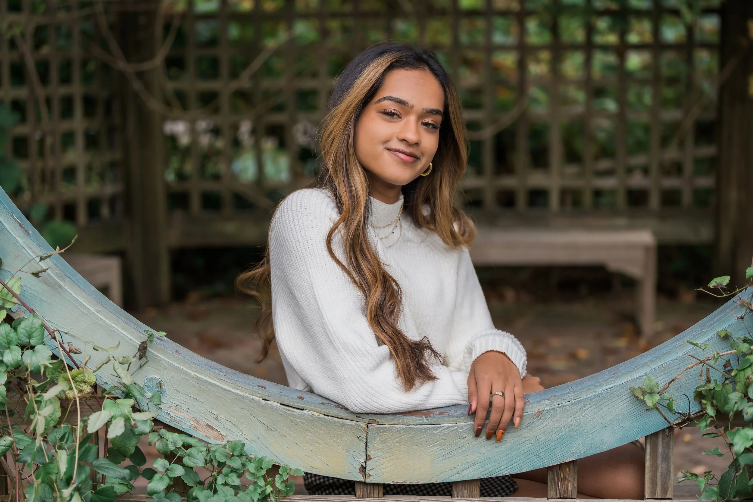 A young woman with long wavy brown hair wearing a white sweater, sitting outdoors on a curved wooden bench, surrounded by greenery and a wooden lattice fence in the background.
