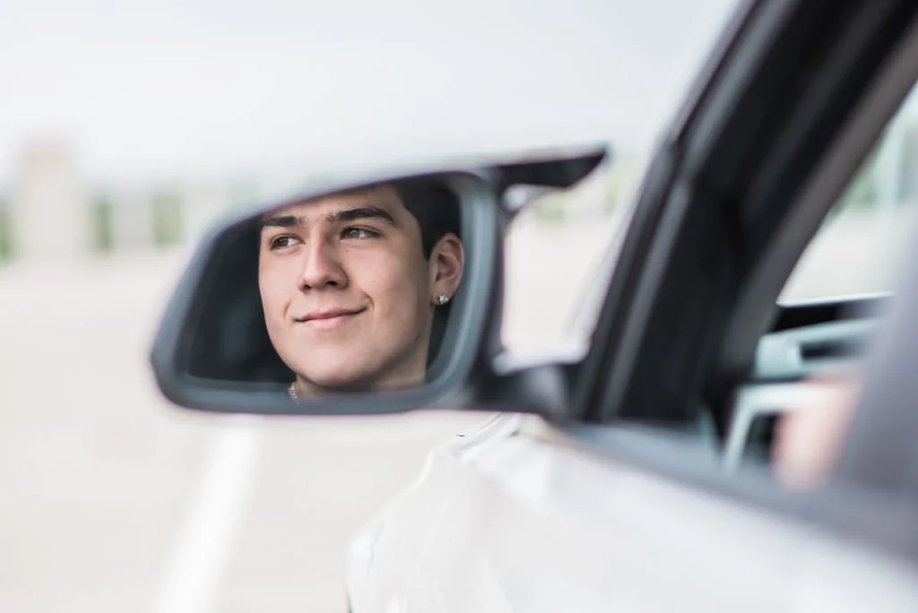 A young man looking at his reflection in a side mirror of a car, smiling with a slight smirk.