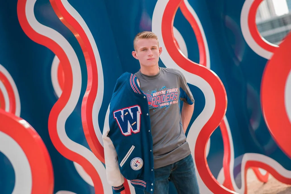 A young man standing in front of colorful, abstract, wavy background art, holding a varsity jacket with a large 'W' patch.