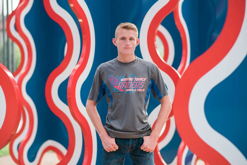 A young man standing in front of an abstract, colorful background with red and white wavy shapes on a blue wall.