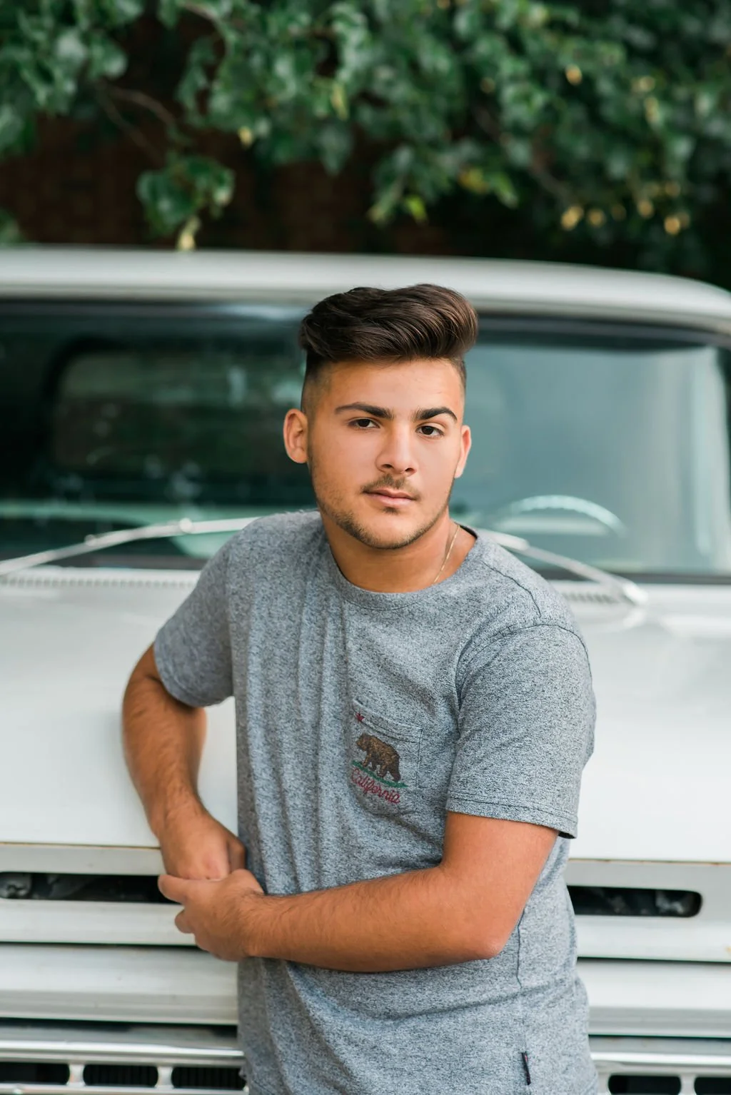 A young man with styled dark hair and a light stubble, wearing a grey T-shirt with a California bear logo, leaning on a white vintage truck, with green trees in the background.