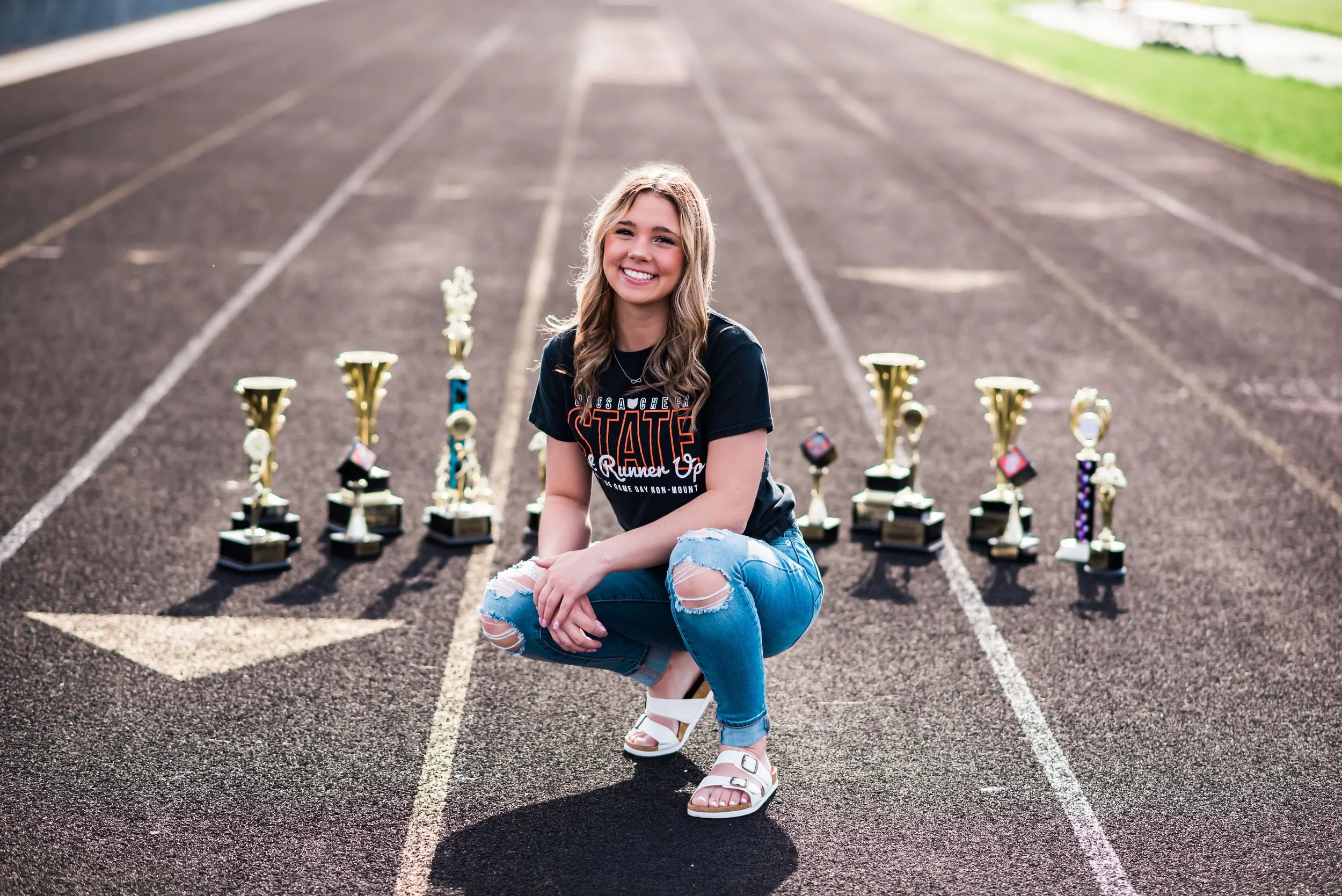 A young woman kneeling on a running track surrounded by multiple trophies, smiling at the camera, wearing a black graphic t-shirt, ripped jeans, and white sandals.