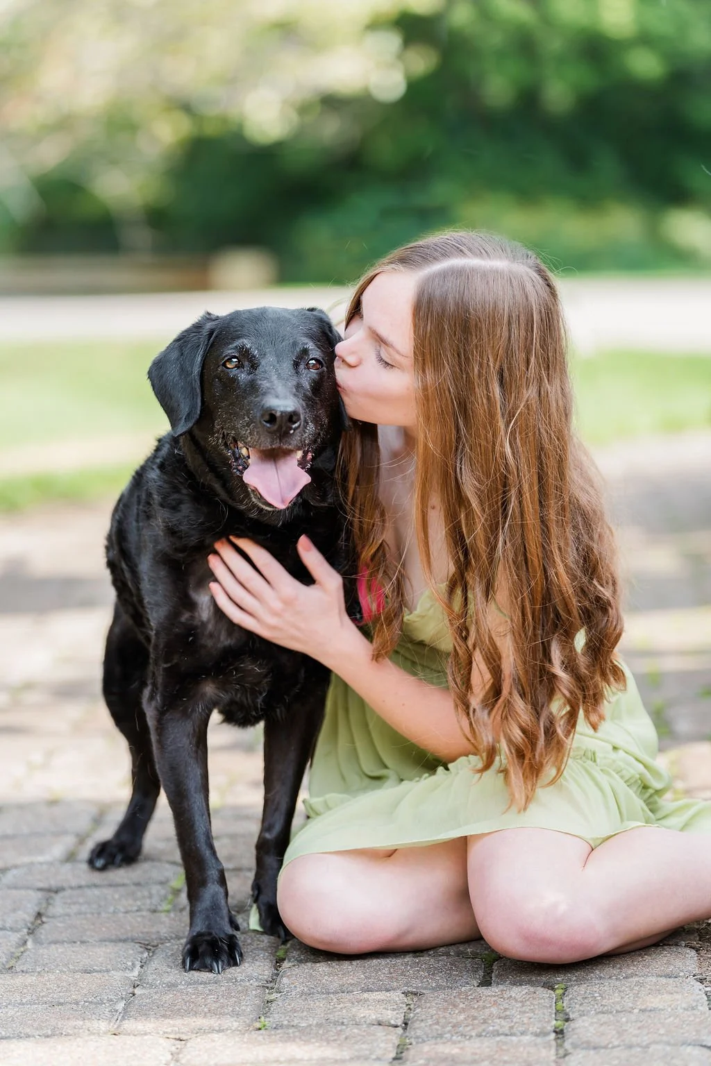 A woman with long reddish-brown hair sitting on a brick pathway, kissing a black Labrador Retriever dog on the side of its face, outdoors with blurred green trees in the background.
