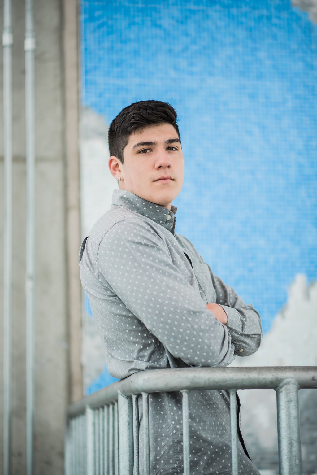 A young man with dark hair and an earring standing with arms crossed in front of a blue tiled wall, wearing a gray, polka-dotted button-up shirt, leaning on a metal railing.