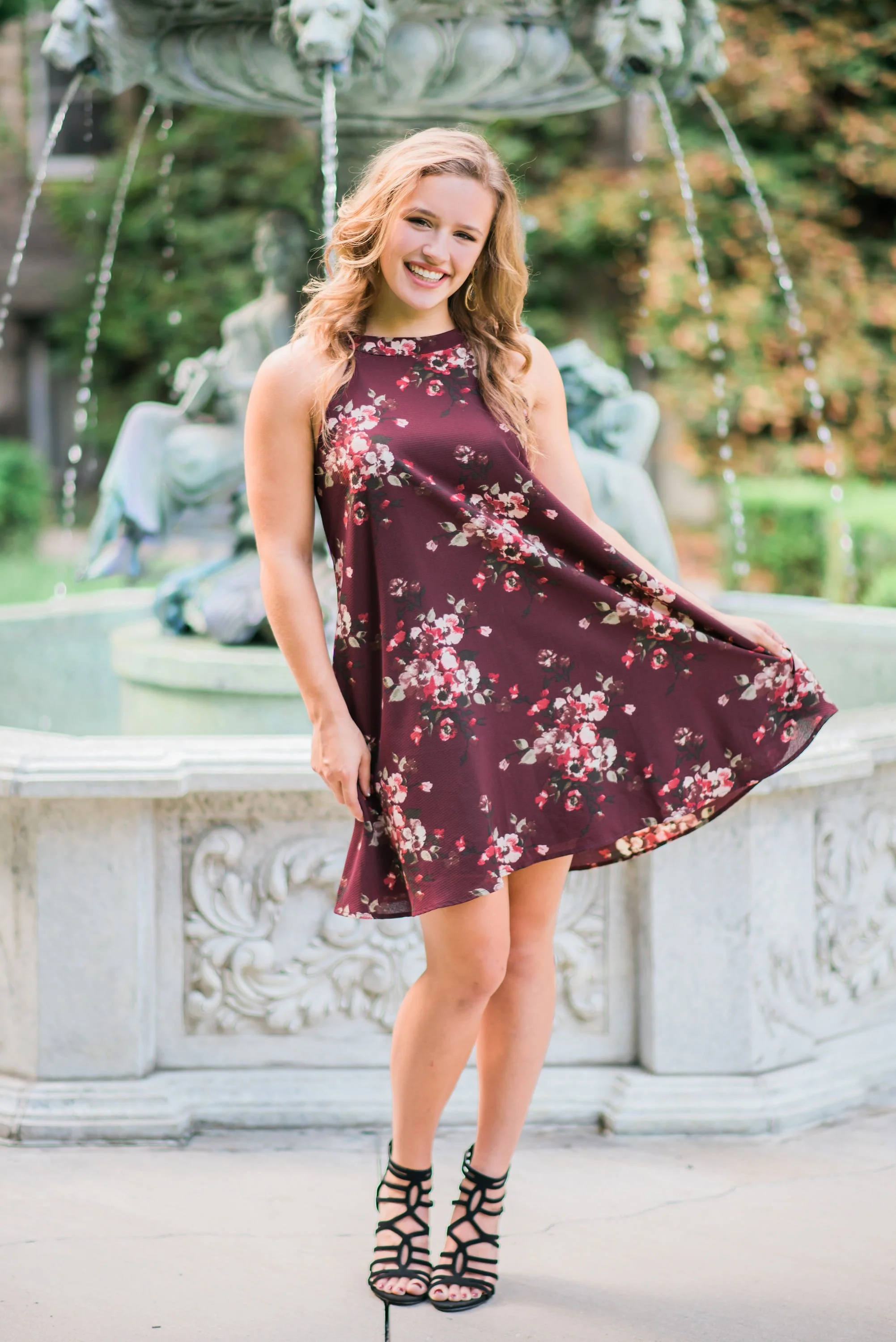 Young woman standing in front of a decorative fountain, smiling, wearing a burgundy floral dress and black strappy high heels.