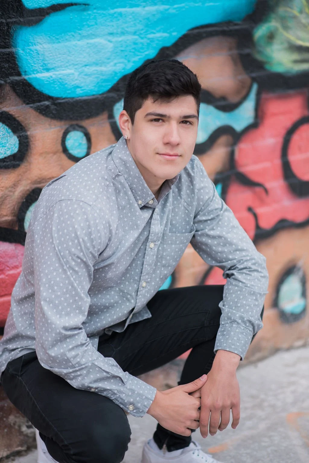 A young man squatting in front of a colorful graffiti mural, wearing a gray polka dot shirt and black pants.