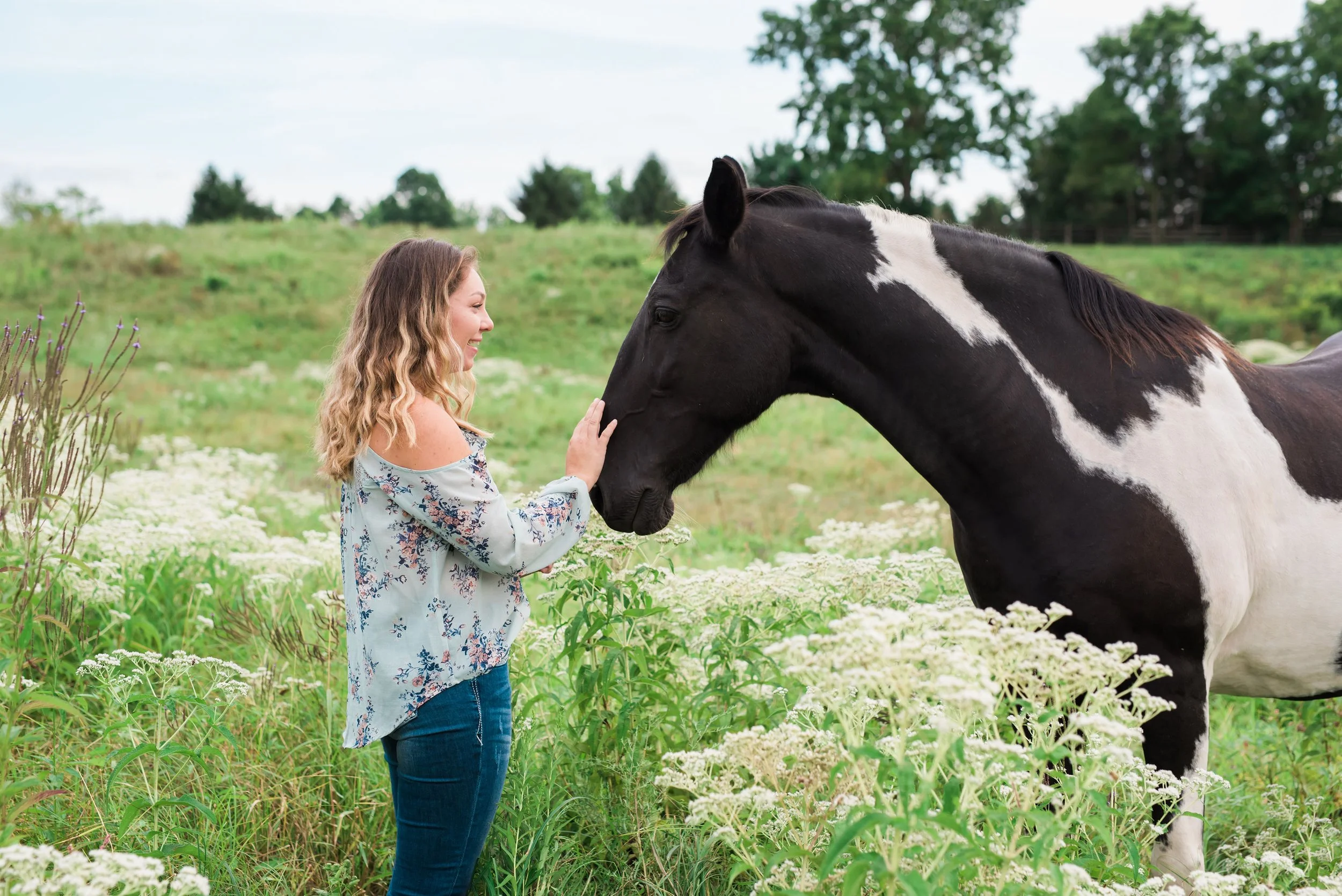 A woman smiling and petting a black and white horse in a green field with wildflowers and trees in the background.