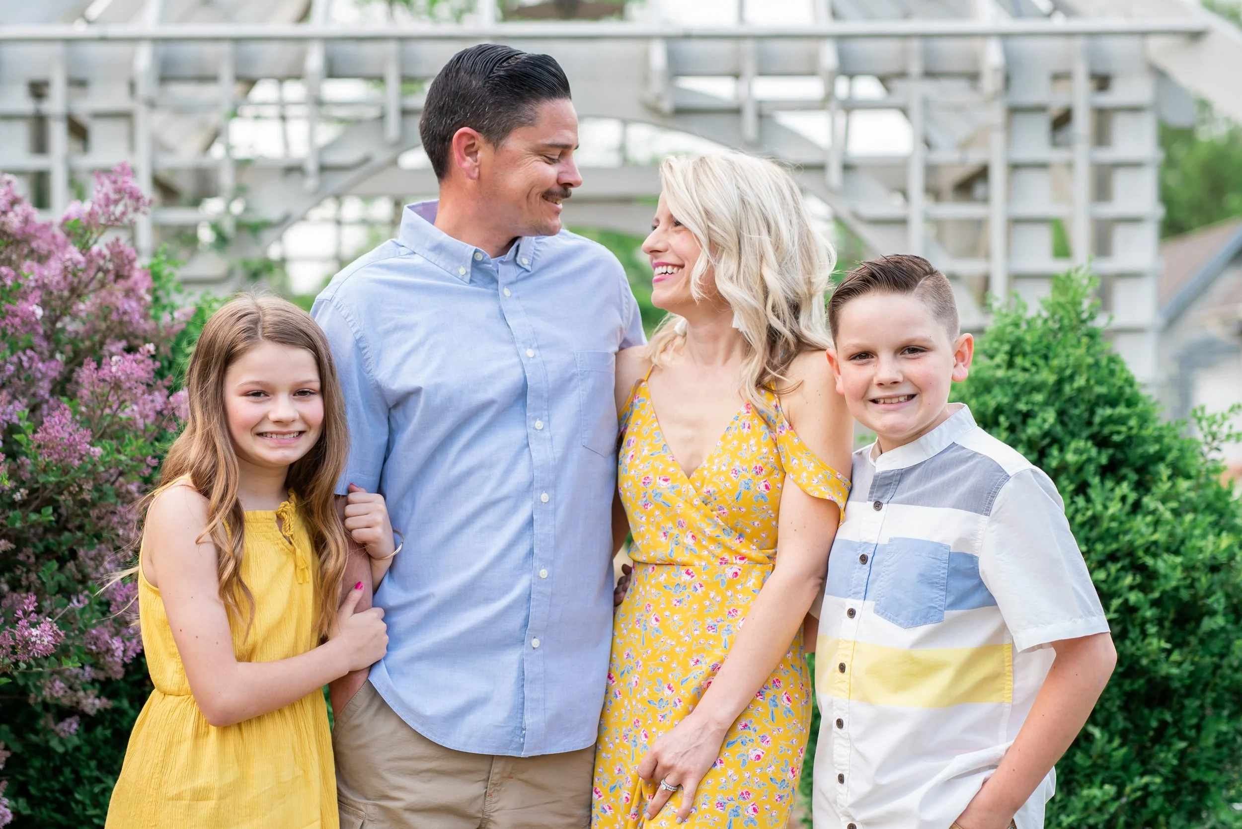 A happy family of four standing outdoors in front of greenery and a house-like structure, smiling and looking at each other.