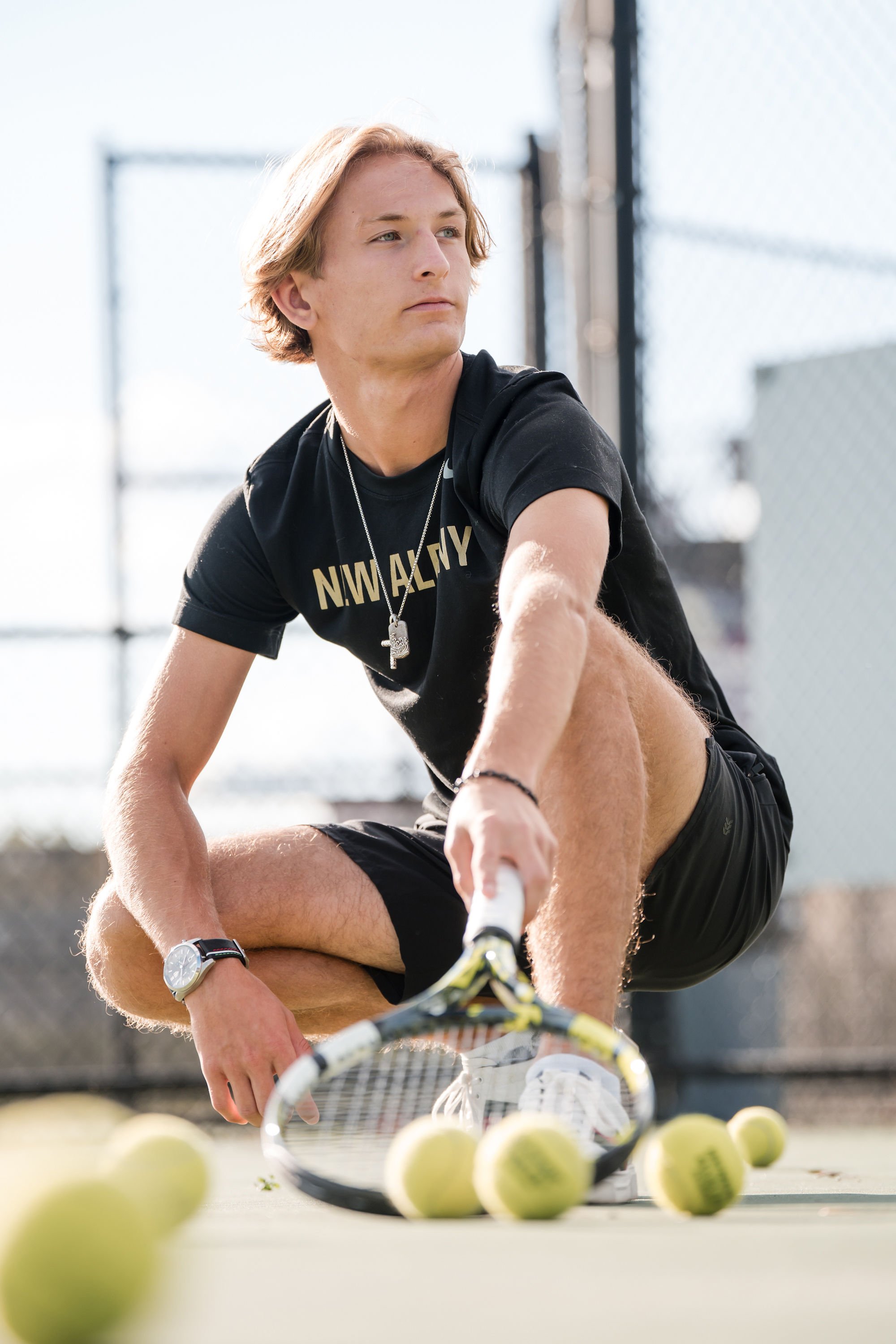 A young man with blonde hair wearing a black T-shirt, black shorts, a wristwatch, and a necklace crouches on a tennis court surrounded by tennis balls, holding a tennis racket.