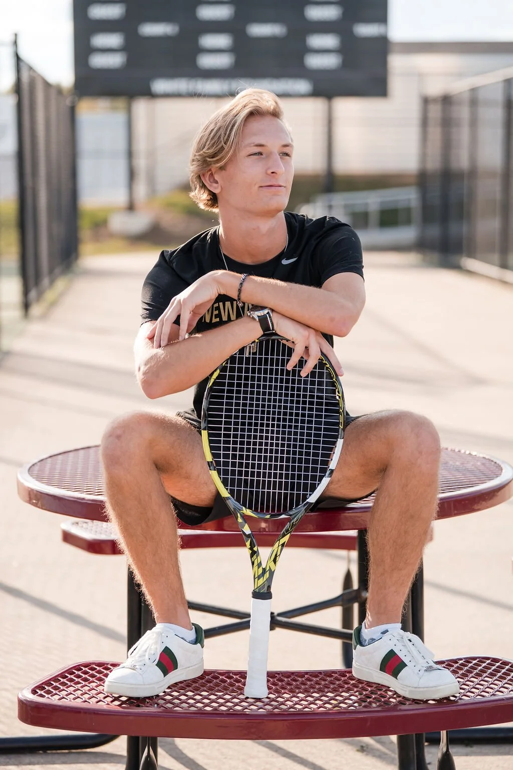 A young man sitting on a red metal table holding a tennis racket with his arms crossed, outdoors at a tennis court, wearing a black t-shirt, white sneakers with green and red stripes, and a smartwatch.