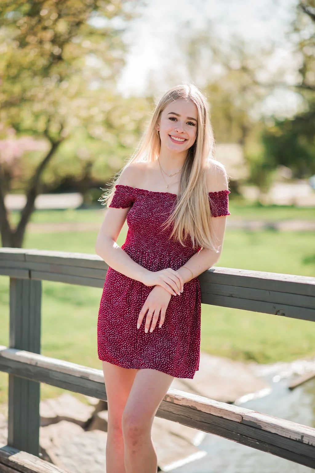 A young woman with long blonde hair smiling, wearing a red off-the-shoulder dress with white dots, standing outdoors by a wooden railing in a park with green trees in the background.