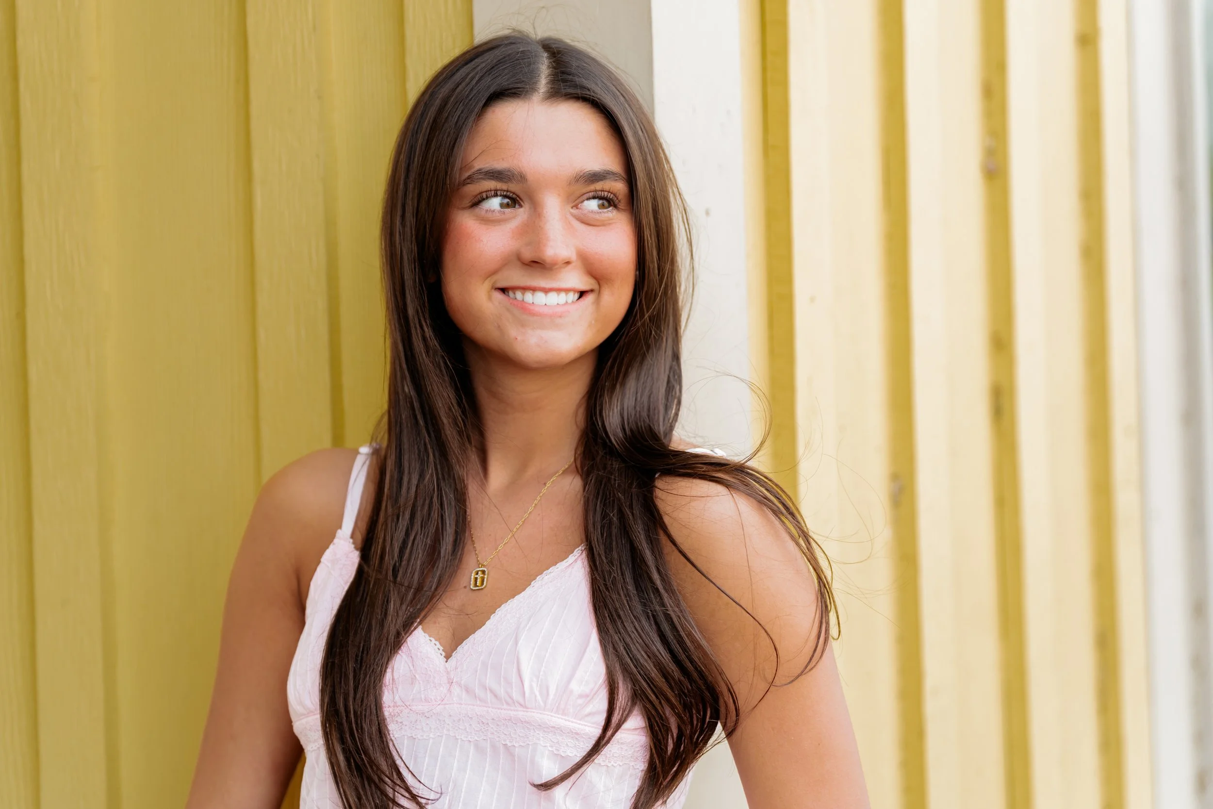 Young woman with long brown hair smiles and looks to her right against a yellow wooden wall.