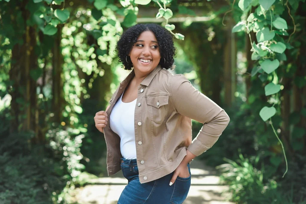 A woman with curly hair smiling outdoors in a green garden, wearing a tan jacket, white shirt, and blue jeans.
